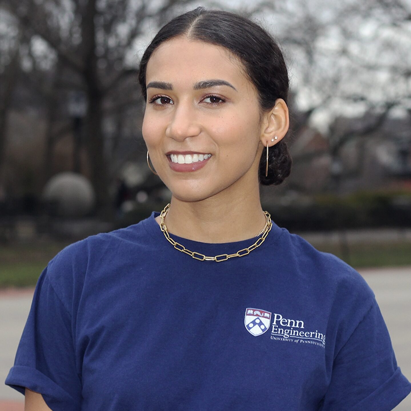 A young woman with dark hair pulled back and smiling. She is wearing a navy Penn Engineering t-shirt and is standing in front of a wooded background. 