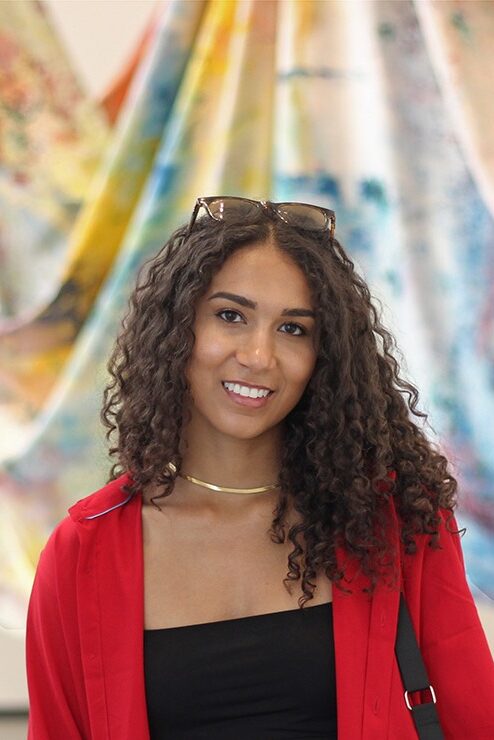 A young woman with dark curly har is posed in front of a draped colorful material background. The woman is wearing a business professional red over jacket, back shirt, and a gold statement necklace.