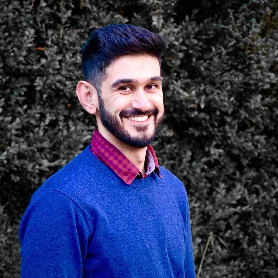 Saad Fahim is young professional man who is dressed in a red collared shirt and a blue pull over sweater. He has short cut dark hair on the sides a full high cut on top. He has a full close cut beard and a wide smile. 