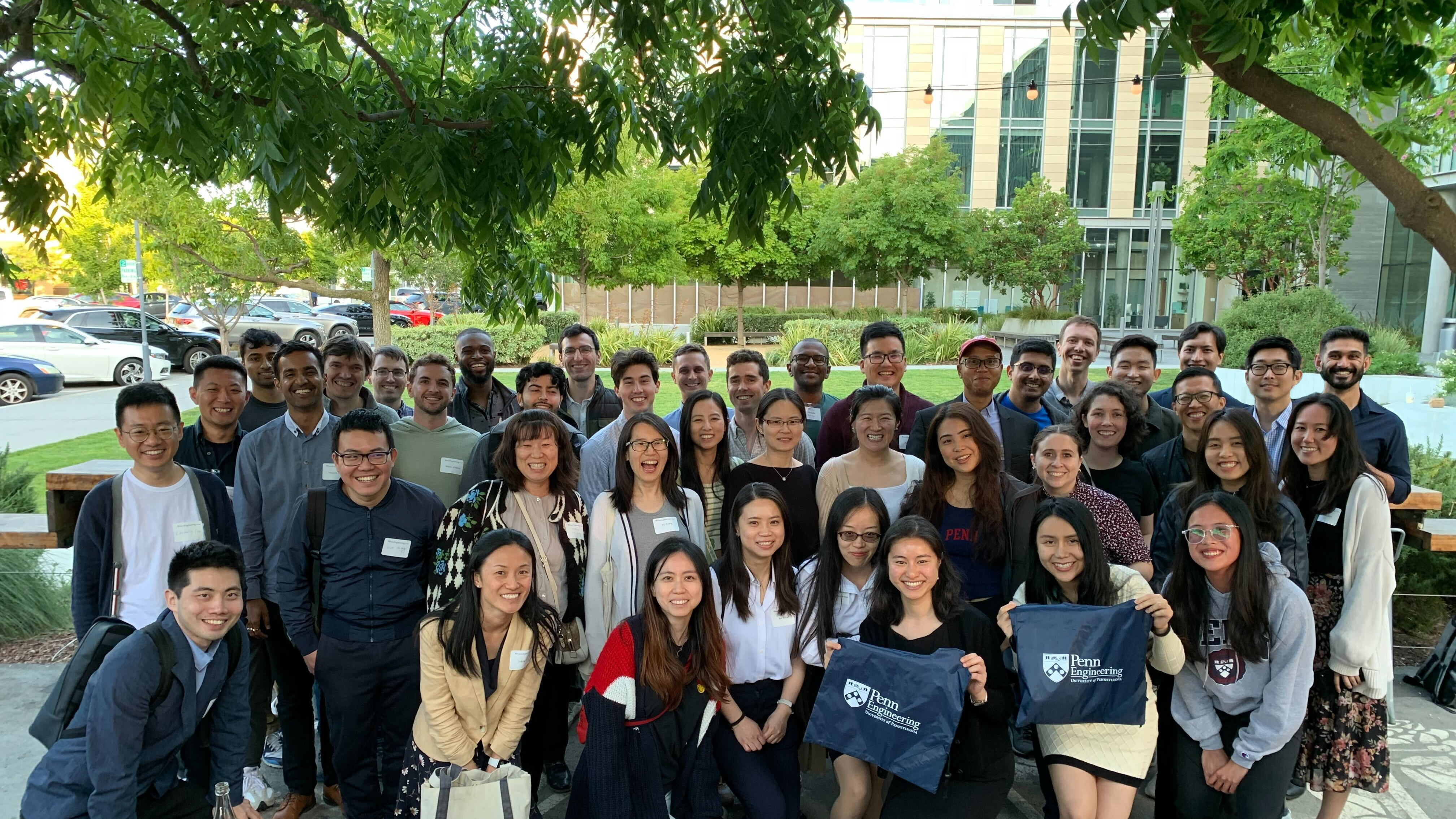 A group of men and women gather around holding up their Penn Swag to show off their school spirit as they gather for an afternoon together outside under the trees in the corporate courtyard. 